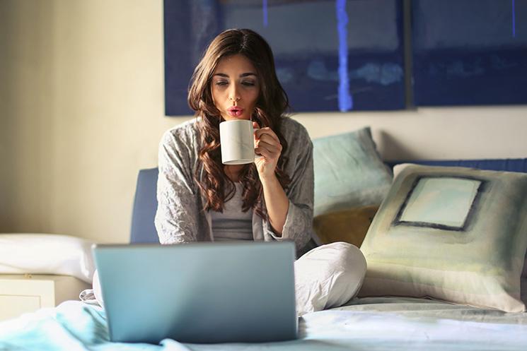 Une femme en train de boire un café et de regarder un ordinateur sur son lit 