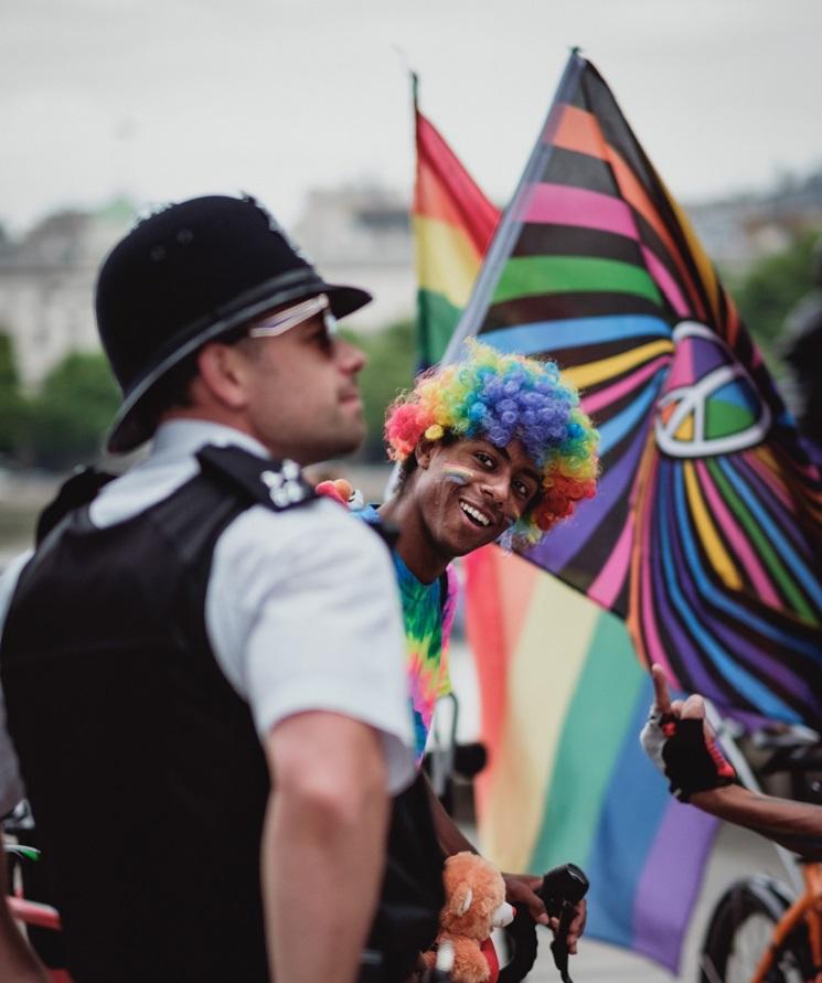 Un policier flouté au premier plan, et derrière lui un homme souriant, les cheveux et le maquillage aux couleurs de l'arc-en-ciel. 