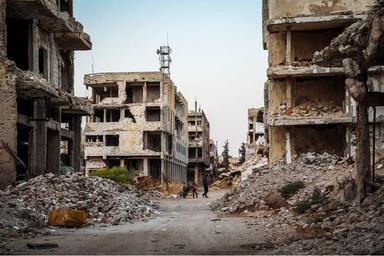 Nombreux débris de bâtiments dans une rue. Deux personnes figurent au milieu des ruines.