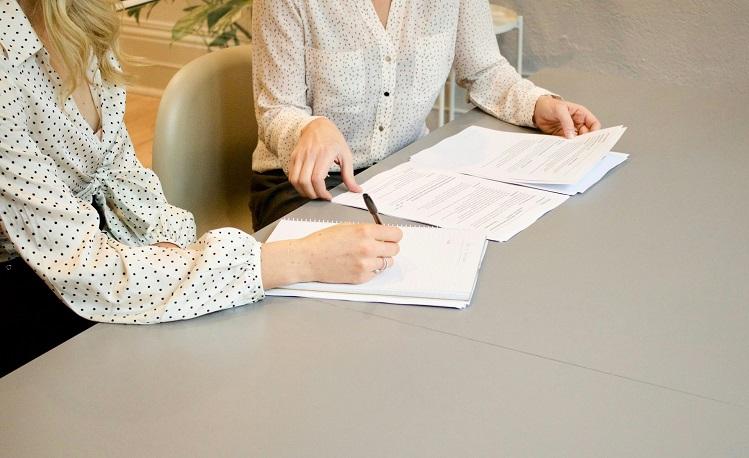 une femme écrit sur une feuille blanche à côté d'une femme qui regarde des documents